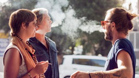 Three people are standing together outdoors - one woman has a cigarette in her hand while the other two men are blowing tobacco smoke out of their mouths