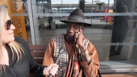 A man in a black trilby hat and tan-coloured jacket and patterned scarf talking to a reporter on a bench. The reporter has a black top and hand-held microphone
