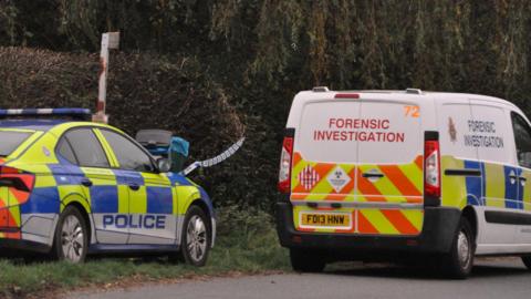 Two police vehicles, a car and forensic investigation van, parked by a hedge and trees with a piece of police cordon tape visible between them