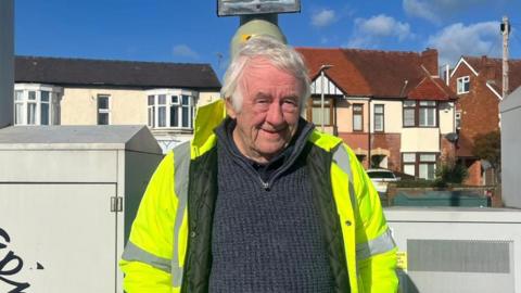 An man with white hair wearing a high visibility coat over a navy blue jumper. He is looking at the camera. Only the top half of his body is visible. You can see a row of houses behind him and a blue sky.