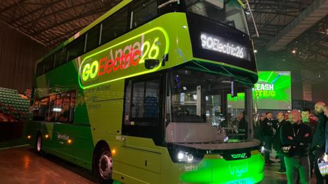 A green double decker bus is parked up, with its lights on, inside a stadium. People are standing around admiring it. The words "Go Electric 26" are projected onto the side.
