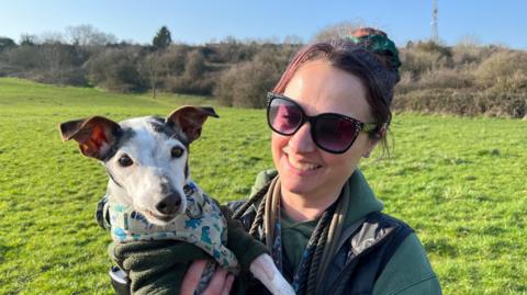 Sophie holding her dog on a sunny day in the park. She is wearing sunglasses and smiling, the dog has a white face and is wearing a bandana.