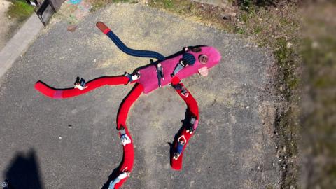An 18-metre long sock monkey lies on the groun. It has a pink body and red arms and legs. There are people lounging all over the monkey in the sun.
