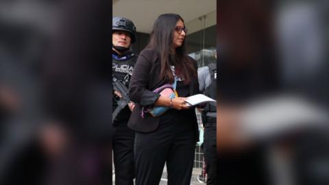 A woman with long dark hair wearing glasses and dressed in black, holds a piece of white paper