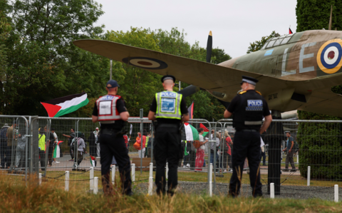 Three male police officers, seen from behind, standing guard as demonstrators take part in a protest behind a wire fence. Some are holding Palestine flags. Between the two groups is a fibreglass model of a vintage aircraft.