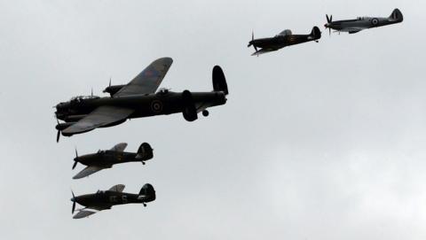 Four aeroplanes are silhouetted against a grey sky. The largest is the Lancaster bomber, and alongside it, are four spitfires.