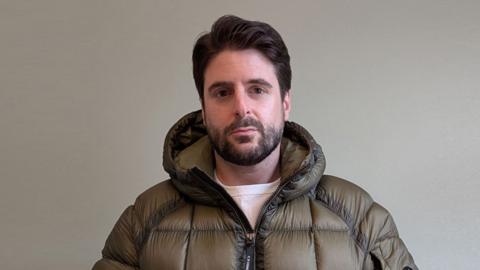 Alex Grossomanides has brown hair, brown eyes and a beard. He is wearing an olive green puffer coat and looking at the camera while standing in front of a white wall indoors.