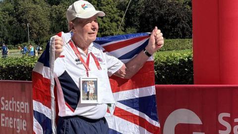 An 89-year-old man wearing Team GB sports uniform and a white cap, holding a union flag behind him, and wearing a photograph of himself and his wife around his neck.