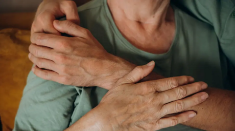 A stock image of an elderly woman, wearing a green shirt, with a hand across them, as if to comfort them.