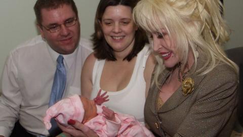 Three adults seated closely together indoors, focusing their attention on a newborn baby wrapped in a pink blanket. In the foreground, Dolly is holding the baby carefully in both hands. She is wearing a light brown checked suit jacket with decorative gold buttons and a flower-shaped brooch on the lapel. The baby is dressed in pink clothing and a matching hat, with one small hand raised upward. To the left of the image, Shane Jarman is seated wearing a white shirt, a light blue tie, and glasses. He appears to be looking down toward the baby. In the centre, Claire is seated wearing a white sleeveless top and is also looking toward the infant.