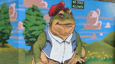 A large section of brick wall that has been painted. It shows green rolling fields and hills with green trees and bushes. The sky is painted blue with white clouds. There is a large green toad wearing a red hat and blue and white shirt with brown shorts.