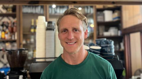 A picture of Drew smiling at the camera. He is wearing a green shirt, has a short beard and blonde hair. He is wearing a green t-shirt. He is stood in front of a coffee machine and a bar.