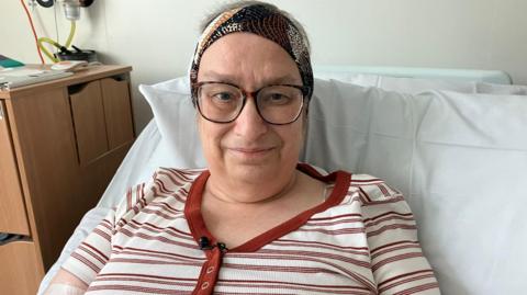A woman in a hospital bed. She is wearing a head scarf and glasses and a stripe white and brown t-shirt. 