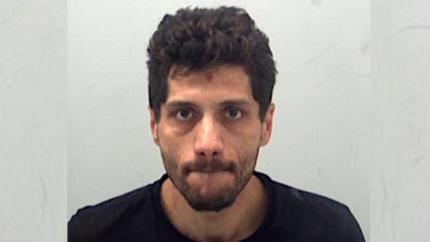 A police custody photo of Jan Drevenak, who has short black hair and a beard. He is wearing a black T-shirt and is standing in front of a grey wall.