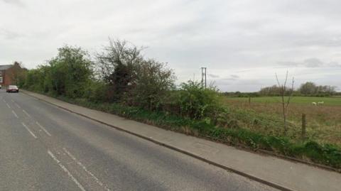 A street view taken on Philadelphia Lane. It is a single carriageway road with a pavement. In the distance is a red car and the start of a row of two-storey brick houses. Behind an unkempt hedge is a field with two white animals grazing.