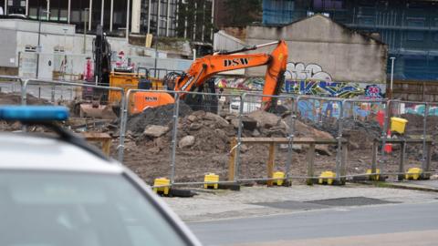 The image shows an orange digger at a construction site cordoned off with metal fencing. A police car is in the foreground. Mounds of earth and rubble are next to the digger.