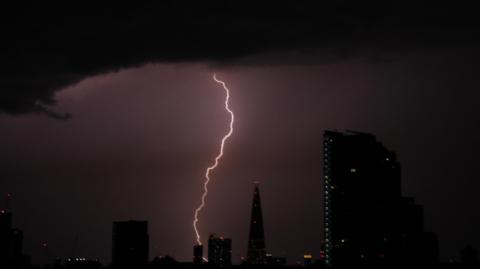 Lightning strikes over a dark London skyline at night, illuminating low storm clouds above silhouetted buildings including a tall glass skyscraper.