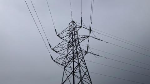 An electricity tower pictured silhouetted against a grey sky in Inverness. The structure is tall with metal lattice work and cables attached to it.