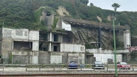 The Summerland site, a derelict building is on a cliff, there is a railway in the foreground and three parked cars.