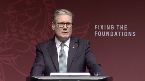 PM Sir Keir Starmer wearing a suit, making a speech at a lectern, standing in front of a backdrop with 'fixing the foundations' written on it 