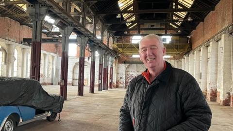 John Kelly stands in the bare red-brick warehouse smiling at the camera. He has short white hair and is wearing a black coat. An old car behind him is partially covered in a black tarpaulin. 