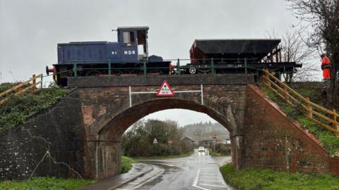 A blue 1959 diesel locomotive pulling a black ballast wagon over a 19th-century brick arch railway bridge above a narrow road. The loco has straight sides with a high cabin and lower front. It is a rainy day and the road is wet. The photographer is at road level and a car is approaching with its lights on in the distance.