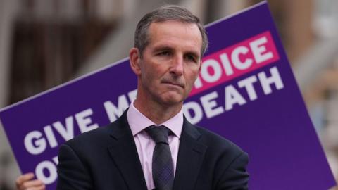A man with a dark suit and dark tie standing before a placard saying give me choice over my death 