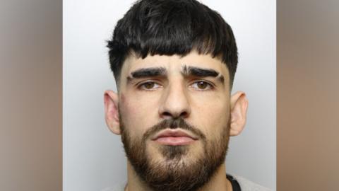 Police mugshot of Leyton Davies. He has black hair and a black beard. 
