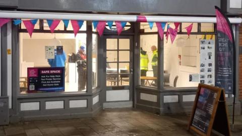 The front of a high street unit which has pink and blue bunting over the door and windows. A sign on the window says "safe space". The light is on inside and several people are standing around.