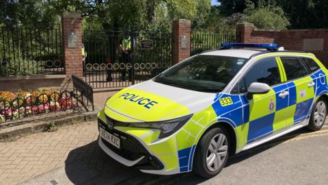 A police car with blue and yellow high-vis decals parked outside the metal gates of Queen's Park in Swindon. There are flowerbeds on either side of the gates and inside there is a paved path bordered with mature trees. Two police officers are standing monitoring the entrance.