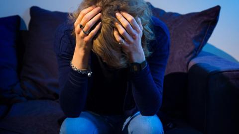 A person with long brown hair sits with their head in their hands showing distress. They are sat on a purple sofa and are wearing a silver bracelet, a navy jumper and jeans.