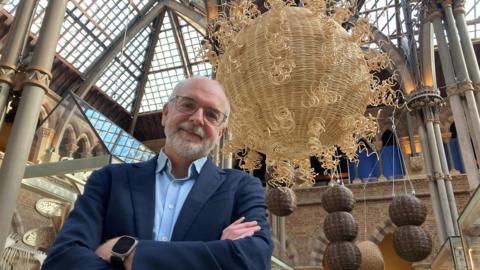 Prof Sir Andrew Pollard from the sternum up - he has his arms crossed and is wearing a blue shirt and blazer. The art installation and museum is behind him.