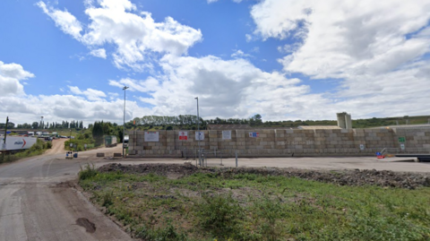 An industrial site with bricked wall and a muddy road leading to it and many lorries parked near it