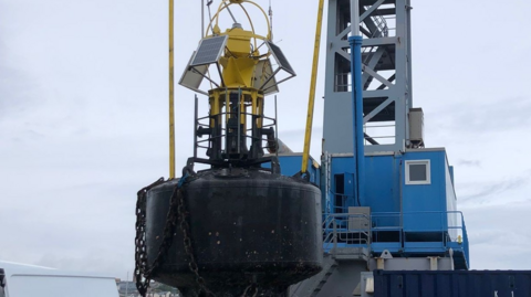 A black and yellow navigational buoy suspended from a crane on a specialist ship