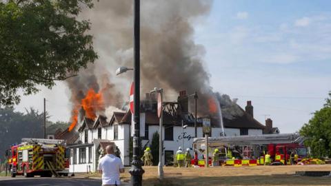 A white pub restaurant building with orange flames on the roof and smoke rising. It is surrounded by firefighters and two fire engines