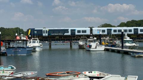 A dark blue and grey SWR train passes over the Lymington viaduct. Small pleasure boats are moored both in front and behind it.