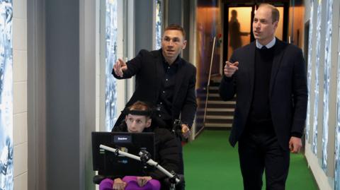 Prince William and Kevin Sinfield accompany Rob Burrow, who is in a wheelchair, down a corridor. They point to something off camera. 