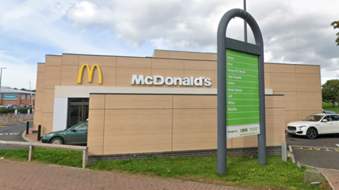 A view of the side of a McDonald's restaurant. There are cars at the drive through, a car park behind, and a green sign showing details of other shops.