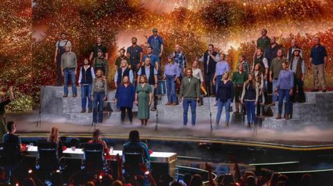 A large group of performers stands on tiered platforms on a brightly lit stage, appearing to sing together as a choir. A conductor stands at the front, guiding the performance. In front of the stage, a panel of judges sits at a table, watching. The background features warm, glowing lights and foliage-like scenery, while an audience in the foreground raises hands and watches the show.