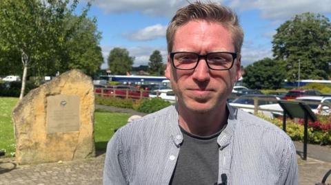 A man wearing a striped black and white shirt, and glasses, stood in a public park.