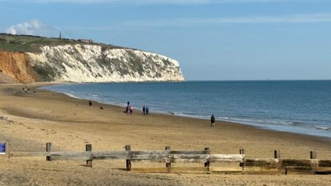 A beach on the Isle of Wight with groynes lining the sand near the front of the image and people walking along next to the sea. There are white cliffs in the distance and blue skies above.