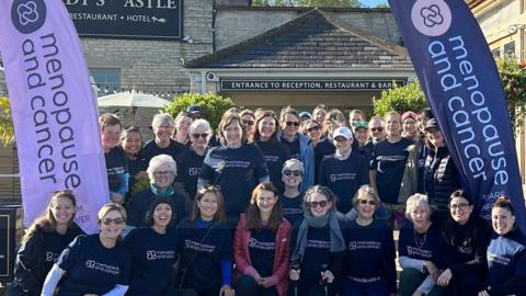 A group of people, wearing dark blue t-shirts with the words Menopause and Cancer. They are posing for a group photograph. There are two banners, one light purple and one navy-blue on each side, with the words Menopause and Cancer. 