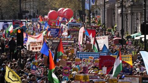 A crowd of people walk London, their heads are seen amongst dozens of colourful signs, flags and plaquards.
