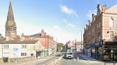 Street view of an urban area with a mix of historic and modern architecture. On the left, there is a tall stone church spire and older brick buildings, while on the right, a row of large red-brick houses and shops.