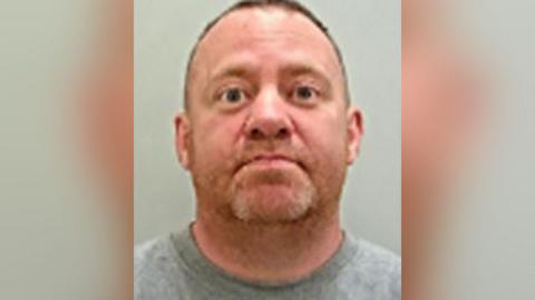 Police custody image of Neil Reid, who has short brown hair and blond stubble. He is wearing a grey t-shirt.