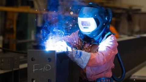 A steel worker, dressed in overalls and wearing a protective mask, welds two sections of a fabricated steel beam at Severfield Plc steel fabricators