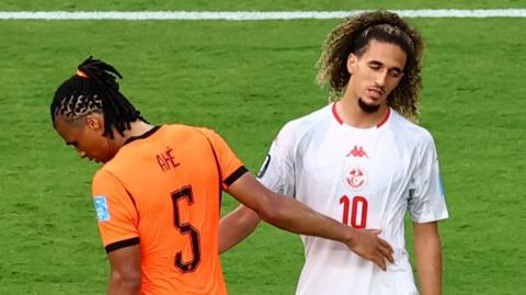 A Netherlands player and a Tunisian player in a half-hearted embrace. The Dutch player on the left is looking down with his back to the camera. The Tunisian is facing the camera and looks dejected.