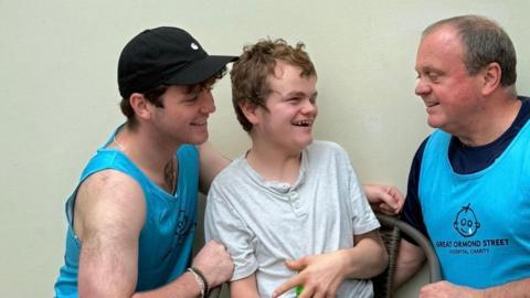 Three men sat together. They are all smiling. The man on the left is wearing a blue vest and black cap and has brown hair. He is looking at the people to the right. In the middle is a younger man, he is looking at his dad on the right. He has light brown hair and is wearing a grey top. On the right is a man looking at the two on his left. He has short hair. He is wearing a black top and blue vest.