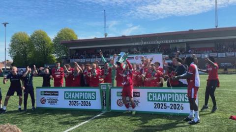 A football team with red kits celebrating and lifting a trophy. They are in front of a sign which says National League South Champions 2025-2026.
