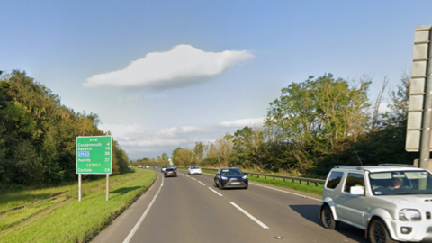 The A66 on a sunny day. It is a single-carriageway road with several cars travelling on it. There is a green road sign listing Cumbrian towns such as Keswick and Cockermouth.